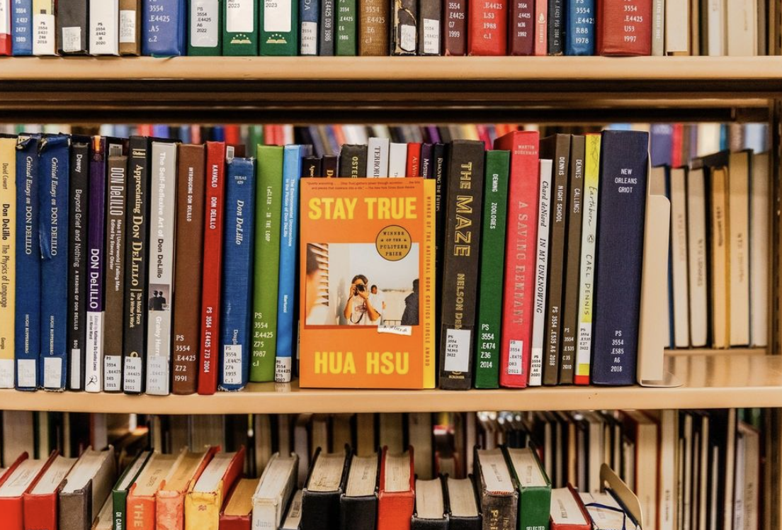 A library bookshelf with an assortment of academic texts and novels. Stay True by Hua Hsu in the center of the shelf with the front cover facing out.